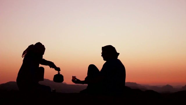 A Bedouin man pours tea and toasts a Western tourist in a silhouette shot.