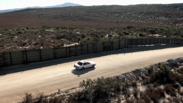 A car drives along a road bordered by a high fence.