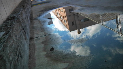 Time lapse of clouds passing over an urban building as reflected in a puddle on the pavement.  - Powered by Adobe