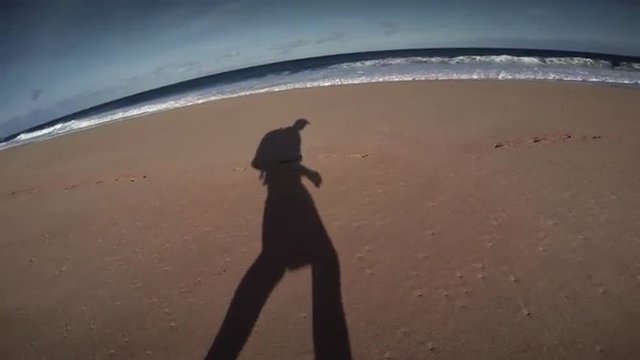 A man's shadow walks along a beach with a backpack.