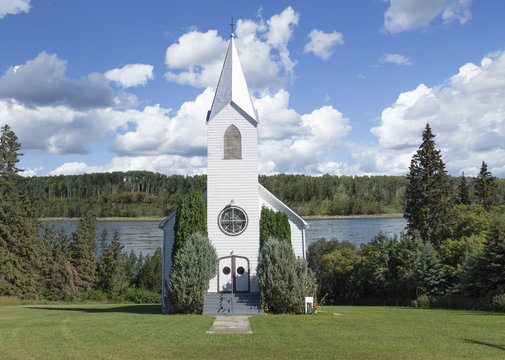 incredibly beautiful image of a white church with steeple sitting on a green lawn flanked by green trees and a big blue lake flowing gently in the background under a blue sky with white puffy clouds.