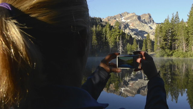 A women taking a photo of a morning reflection of the Sierra Buttes on Sand Pond in Tahoe National Forest, California.