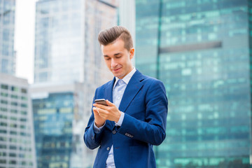 Fototapeta premium Handsome businessman in suit standing at the street with smart phone in hand. Modern skyscrapers on the background.