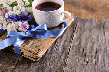 Old book with beautiful flowers and cup of tea on wooden table close up