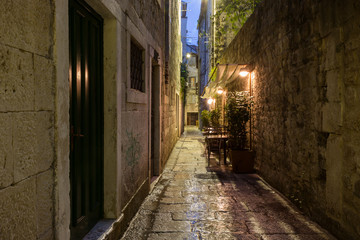 Fototapeta na wymiar Narrow and empty alley with few cafe tables at the old town in Split, Croatia, at dark after rain.