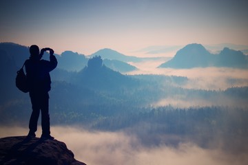 Fototapeta na wymiar Tourist with backpack takes photos with smart phone on peak of rock. Dreamy fogy valley below