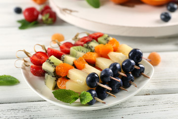 Fresh fruits on skewers in plate on wooden table, closeup