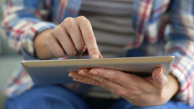Tilt up shot of female hands using tablet PC. Midsection of woman in casuals is scrolling and zooming. She is sitting on sofa in brightly lit house.
