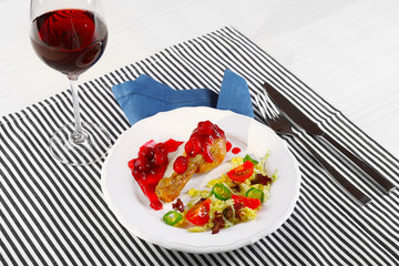 Dish of baked chicken leg and vegetable salad in white plate with glass of wine on table with napkin, closeup