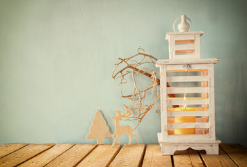 white wooden vintage lantern with burning candle, wooden deer, christmas gifts and tree branches on wooden table. retro filtered image
