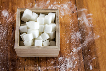 Sugar Cubes in Square Shaped Bowl with Unrefined Sugar spill over in Wooden Background