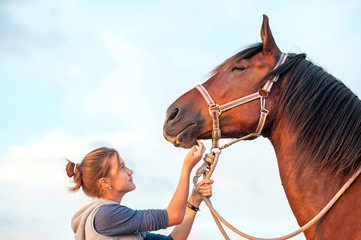 Young cheerful teenage girl stroking brown horse's nose. Outdoor