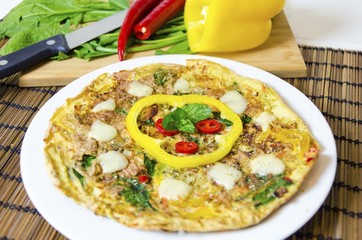 A spinach, chili, mint and pepper omelette in a dish over bamboo table cloth and chopping board with knife and raw vegetables on while background. A healthy diet meal omelet.