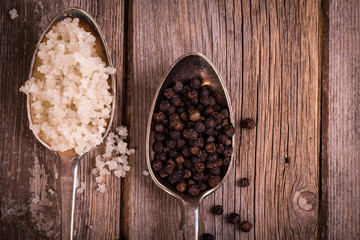 Vintage effect image of tarnished silver spoons filled with salt crystals and black peppercorns, over rough wood background. Space for your text