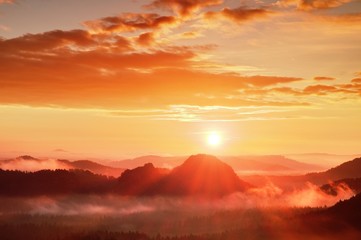 Red misty daybreak. Foggy autumn morning in a beautiful hills. Peaks of hills are sticking out from rich colorful clouds.