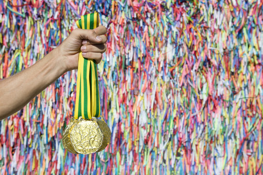 Hand of Brazilian athlete holding gold medals in front of a colorful wall of wish ribbons at the Bonfim church