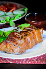 Grilled beef steak with salad and sauce on wooden table at white plate. Salad with tomatoes and cucumbers and sauce barbecue. Leafs of basil as decoration.