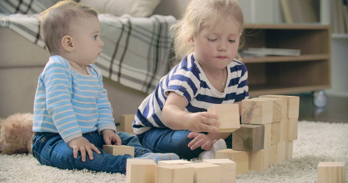 Adorable baby boy sitting next to his sister stacking toy blocks 