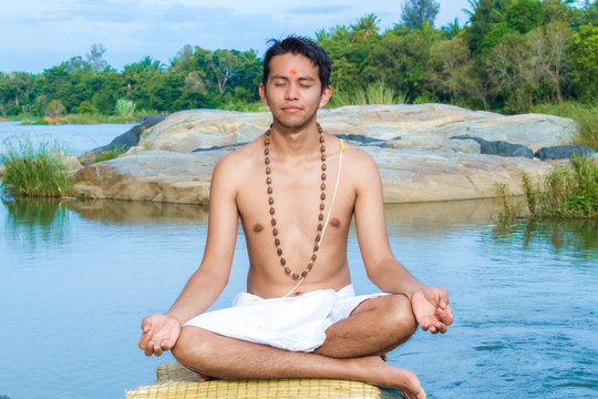 A young brahmin sits in meditation on a river bank in India 
