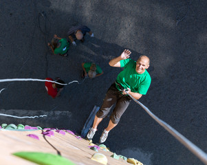 Smiling Man Descending on Rope from Top of Climbing Wall Positive Mature Extreme Climber Greeting Waving Hand Hanging High on Belaying Rope on Outdoor Rock Climb Gym