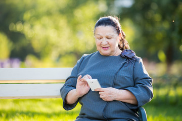 Mature woman using smartphone sitting on a bench in the park. Smiling and touching the screen