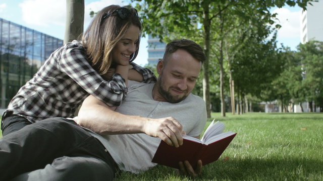 Young romantic couple spending time in park and reading book. Young couple lying on grass and reading book. Happy couple preparing for exams in park.