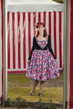 View of pinup young woman in vintage style clothing next to red and white striped tents.