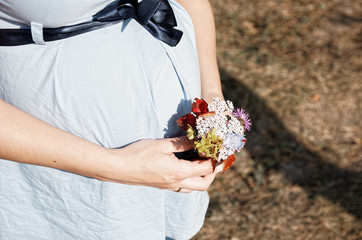 Young pregnant woman in field with a bunch of wild flowers under belly. Selective focus.