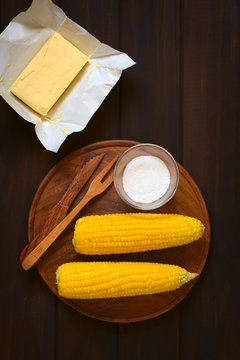 Overhead Shot Of Two Cobs Of Cooked Sweet Corn On Wooden Plate With Salt And Cutlery, With A Piece Of Butter Above, Photographed On Dark Wood With Natural Light