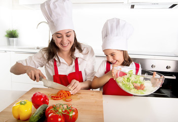 young mother and little daughter at house kitchen preparing salad for lunch wearing apron and cook hat