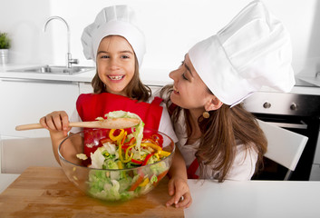 happy mother and little daughter at home kitchen preparing salad in apron and cook hat