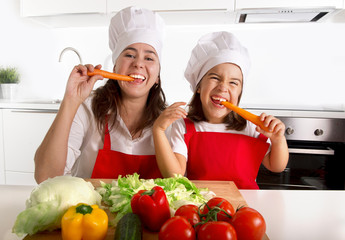 happy mother and little daughter in apron and cook hat eating carrots together 