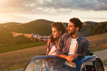 A loving couple, on a summer afternoon, watching sunset from the roof of an old car, around the classical landscape of Tuscany, vineyards and farmland. Woman points to look at a point to her boyfriend - obrazy, fototapety, plakaty