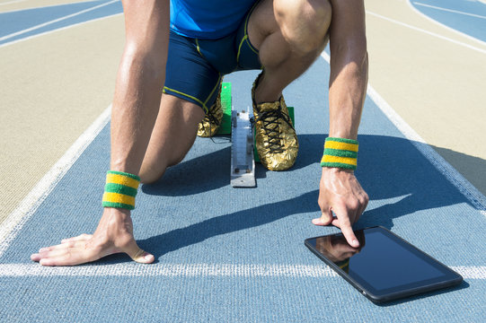 Athlete crouching at the starting blocks of a running track wearing Brazil colors wristbands using his tablet