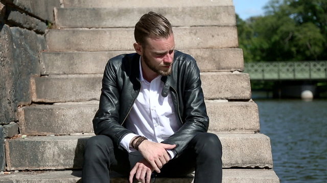 A man in his 20s sitting down on steps outside wearing a white shirt and black leather jacket on a sunny summer day.