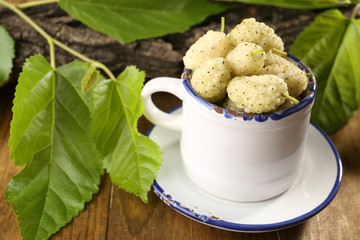 Ripe mulberries in mug with green leaves on table close up