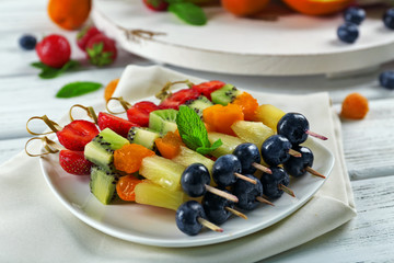 Fresh fruits on skewers in plate on wooden table, closeup