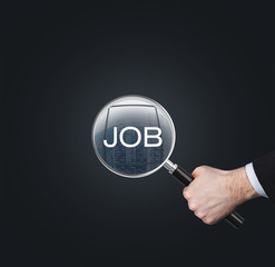 A hand holds a magnifying glass with the word 'JOB'. The concept of the searching of the job. Dark background.