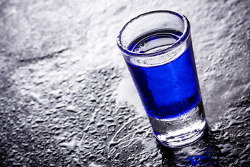 Blue liquid in vodka shot glass on black wet surface.