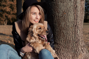 Beautiful cute happy girl in a black hat playing with her dog in a park in autumn another sunny day