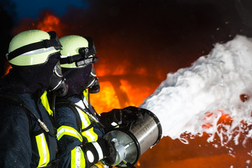 Fototapeta premium Feuerwehr - Feuerwehrmänner löschen ein großes Feuer, sie stehen vor einer Feuerwand und tragen Schutzkleidung