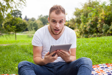 Obraz premium Bearded young man lying in park on blanket. He is using white tablet pc.