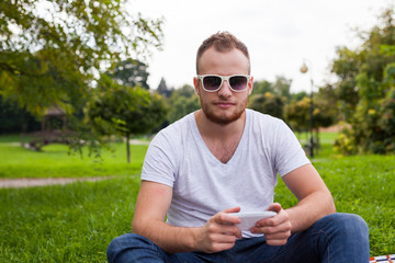 Fototapeta premium Bearded man sitting in park on blanket. He is using mobile phone.