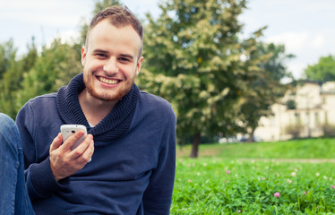 Bearded man sitting in park on blanket. He is using mobile phone.