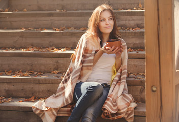 Young woman resting and drinking tea sitting in autumn garden on the steps, wrapped in a woolen plaid blanket.