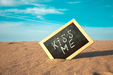 closeup of a chalkboard with a wooden frame and the word kiss me written in it, placed on the sand of a beach