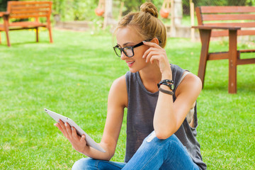 Sensual blonde woman sitting in park on blanket. She is using white tablet pc.