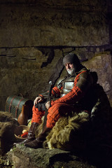 Old medieval King in armor with sword is sitting on furs near th