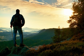 Fototapeta premium Silhouette of tourist with backpack. Sunny spring daybreak in rocky mountains. Hiker with sporty backpack stand on rocky view point above misty valley.