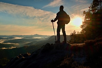 Silhouette of alone hiker with poles in hand. Tourist with sporty backpack stand on rocky view point above misty valley. Sunny spring daybreak in rocky mountains.